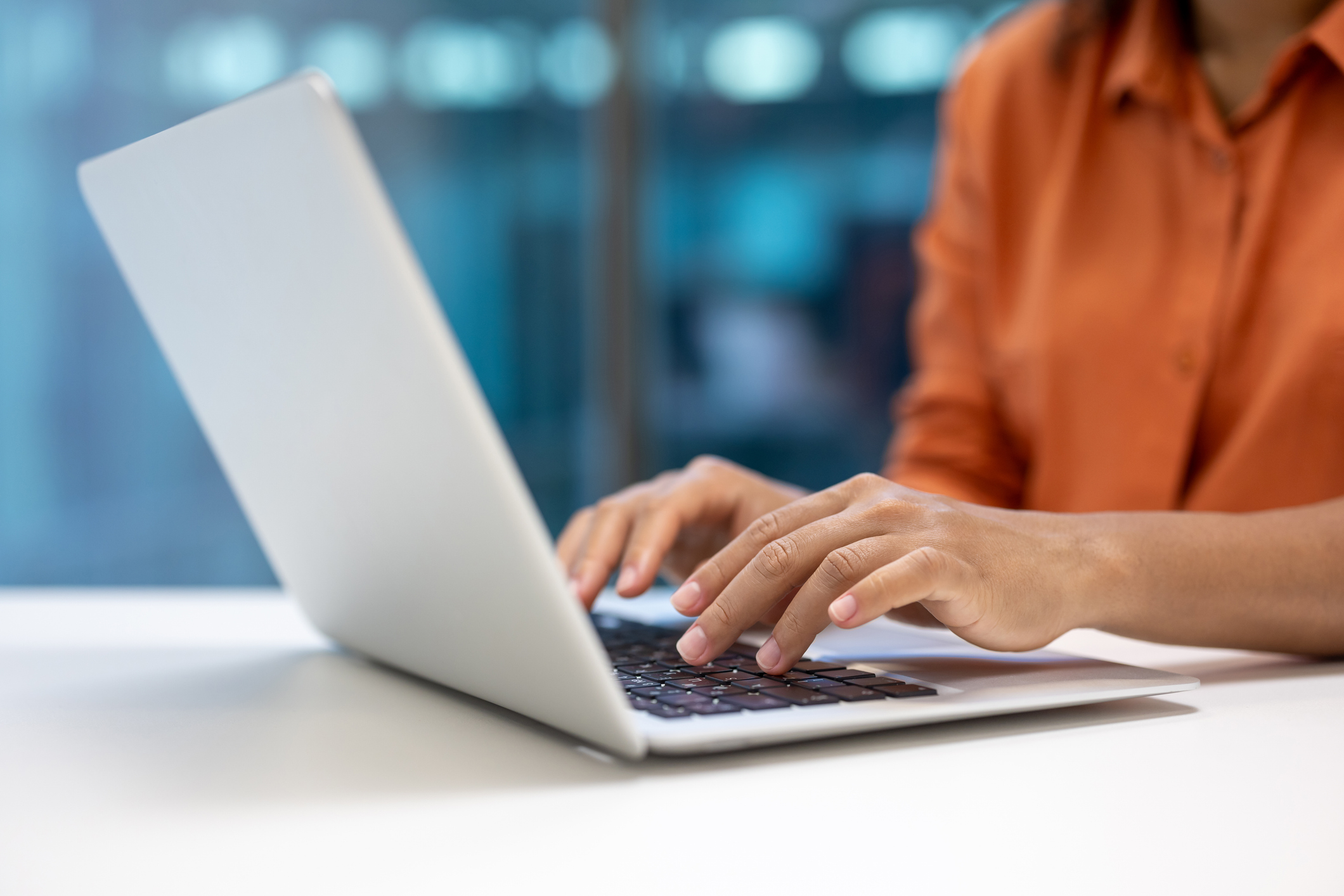 Hands typing on laptop in office. Business professional in orange shirt working efficiently. Captures modern office atmosphere, digital communication, and professional work culture.